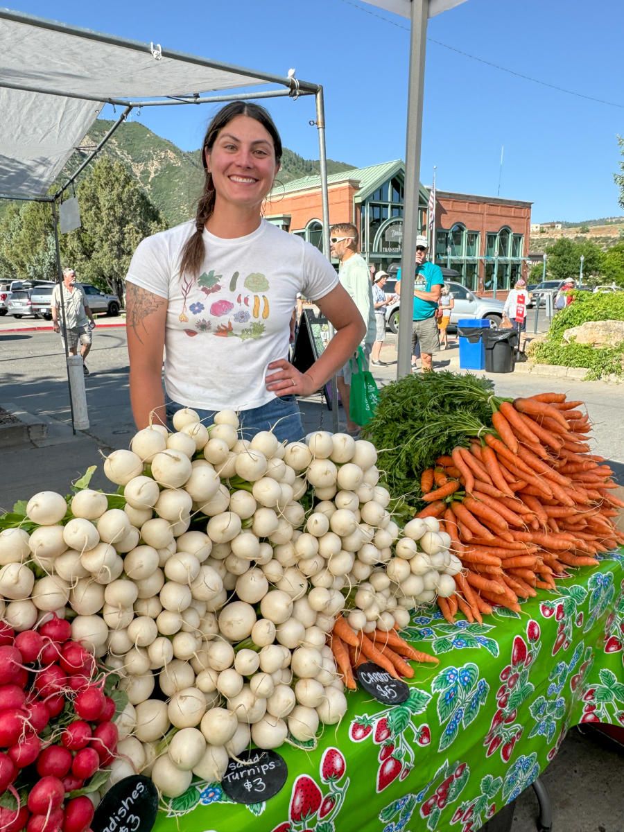 A smiling seller standing behind a huge stack of carrots and onions at the Durango Farmer's Market